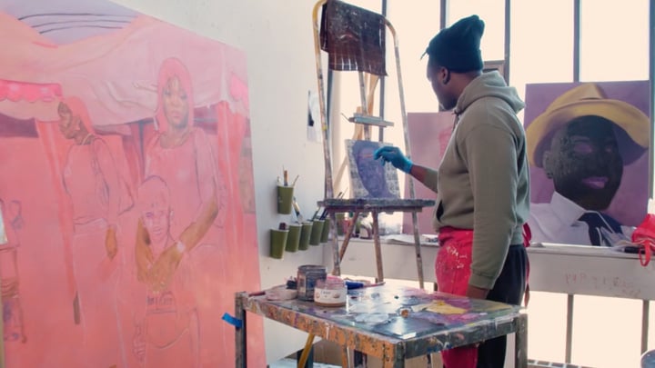 A black student wearing a beanie and gray hoodie is painting a small portrait with a large pink-hued work and bold painting in the background.