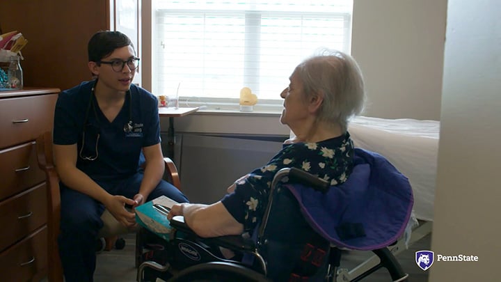 A young nursing student dressed in blue scrubs talks to a patient in a wheelchair.