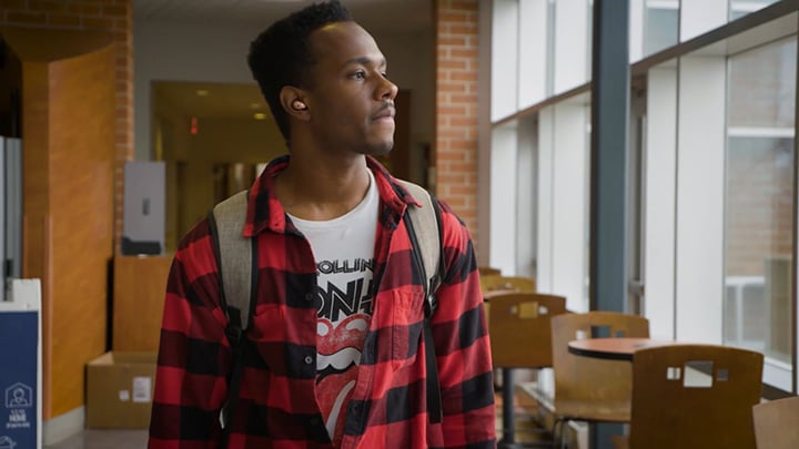 A student wearing a red and black flannel shirt and carrying a backpack walks by a wall of windows.
